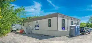 Portable office building with porta potties on a sunny day, set among trees with clear blue sky.